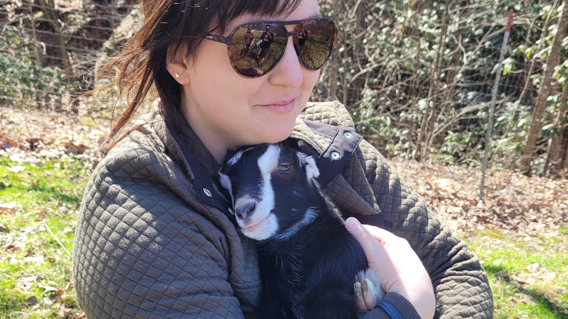Woman in sunglasses cradles baby goat during goat yoga session at farm in Asheville, NC.