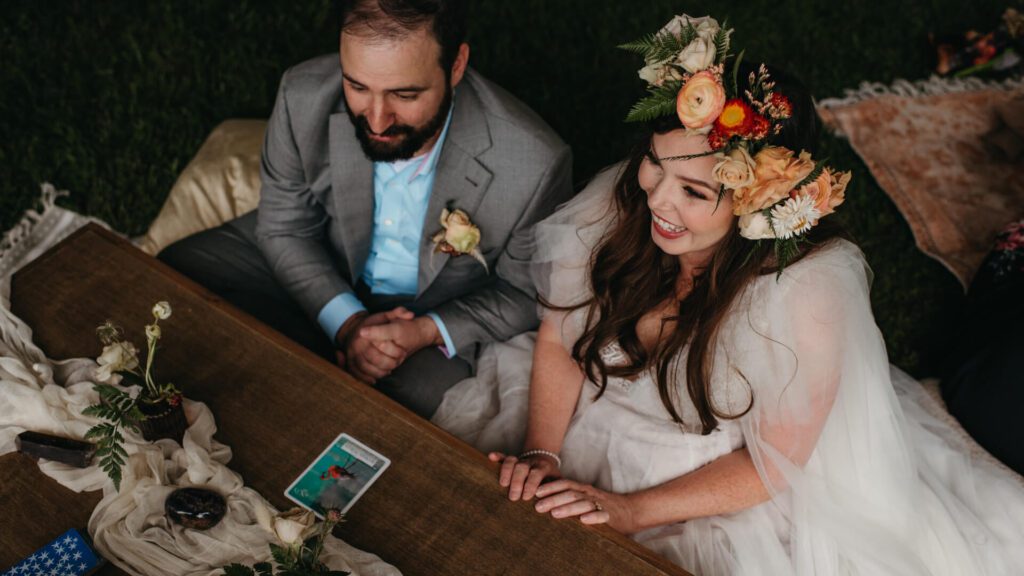 A bride and groom laugh while receiving a tarot reading at their wedding reception. The bride is wearing a lovely flower crown and the mood is light.